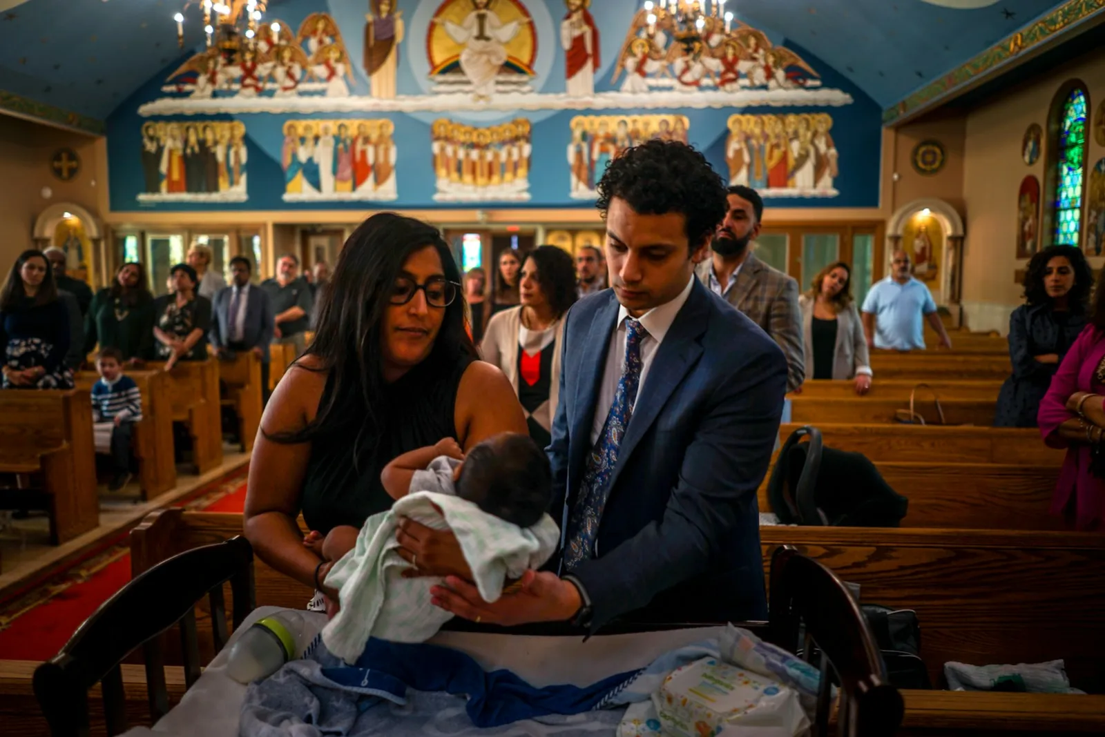 A young couple holding their baby during a baptism, with relatives standing in the church pews behind them